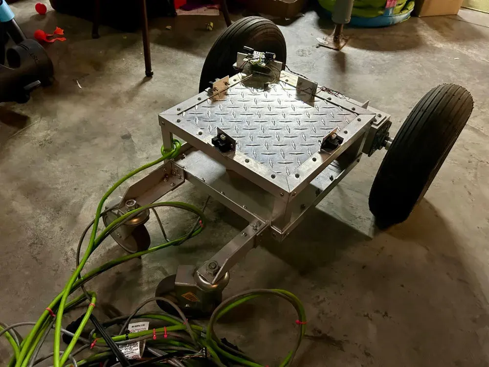 A small 4 wheeled structure made of aluminum angle and diamond plate. There are two large pneumatic back wheels and two small front casters. Sonar sensors are seen on each of the four sides of the top. Extension cords are attached. The robot is sitting in a basement.